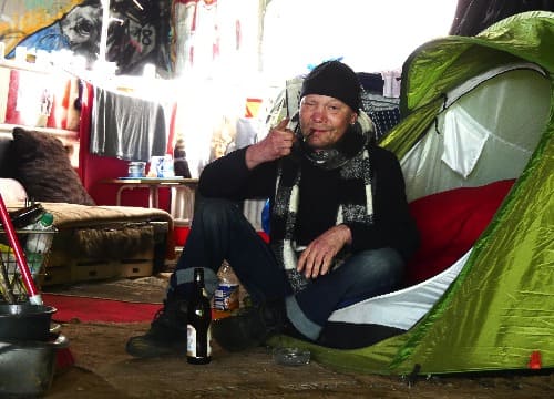 Auf dem Foto sitzt Björn vor seinem Zelt auf dem Boden und raucht eine Zigarette. Im Hintergrund ist seine selbst eingerichtete Ecke zu sehen.