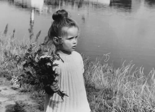 Auf dem schwarz-weiß Foto ist ein Kleinkind, Claudia, vor einem Fluss zu sehen. In der rechten Hand hält sie einen Blumenstrauß.