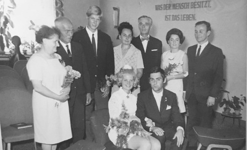 Auf dem schwarz-weiß Foto ist ein Hochzeitspaar, Mann und Frau, in einem Raum zu sehen. Hinter ihnen sind weitere Personen zu erkennen. Die Personen im Raum lächeln. Rechts auf dem Bild ist an einer Wand die Inschrift: „Was der Mensch besitzt, ist das Leben.“ angebracht.