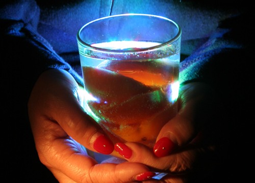Auf dem Foto zu sehen ist ein gefülltes Glas, gehalten von beiden Händen einer Frau. Das Glas ist mit einer Frucht und Wasser randvoll gefüllt.